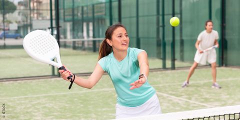 Frau spielt Padel-Tennis auf einem Außenplatz und bereitet sich darauf vor, einen Ball mit einem Schläger zu schlagen. Im Hintergrund ist eine weitere Spielerin zu sehen. Der Text lautet: „TRENDSPORT PADEL-TENNIS.“.