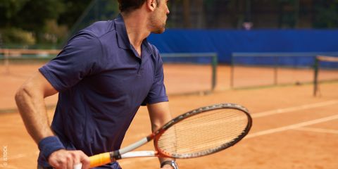 Ein Mann im blauen Sporthemd spielt Tennis auf einem Sandplatz. Er hält einen Schläger in der Hand und blickt zur Seite. Der Text auf dem Bild lautet „TENNISARM ADÉ“. Im Hintergrund sind ein Netz und ein weiterer Platz zu sehen.