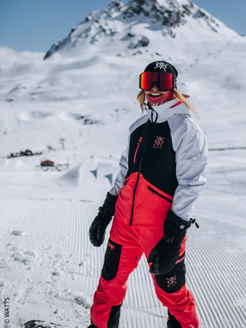 Eine Person in einem rot-schwarzen Schneeanzug steht lächelnd auf einem verschneiten Berg und trägt eine Skibrille, Handschuhe und eine Mütze an einem sonnigen Wintertag.