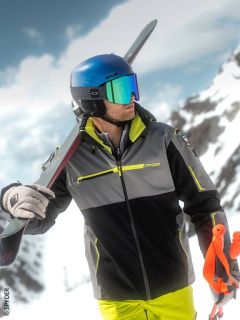 Ein Skifahrer mit blauem Helm und Reflexionsbrille steht auf einem verschneiten Berg, trägt Skier über der Schulter und hält orangefarbene Skistöcke.