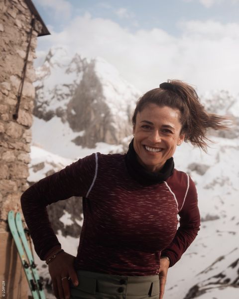 Eine lächelnde Frau in einem kastanienbraunen Oberteil steht im Freien in der Nähe eines steinernen Gebäudes mit verschneiten Bergen und Skiern im Hintergrund.