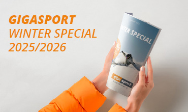 Hände in einer orangefarbenen Jacke halten ein "Gigasport Winter Special 2025/2026" Magazin mit Bergen auf dem Cover vor einem hellen Hintergrund.