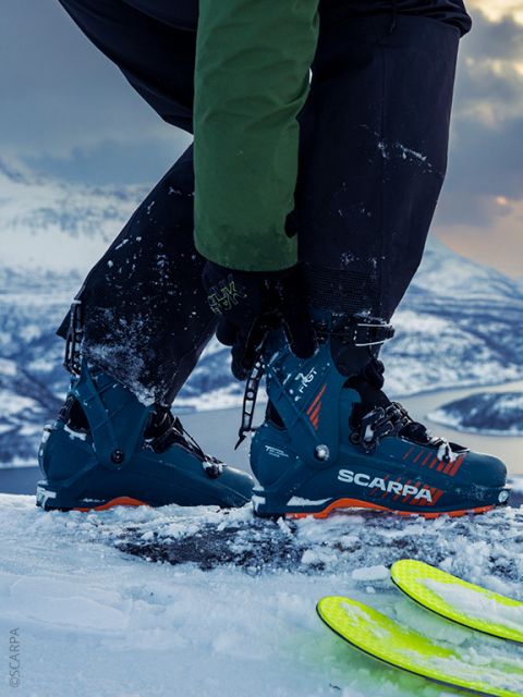 Eine Person, die SCARPA-Skischuhe trägt, stellt ihre Schuhclips auf verschneitem Boden ein, während im Hintergrund Berge und Skier zu sehen sind.
