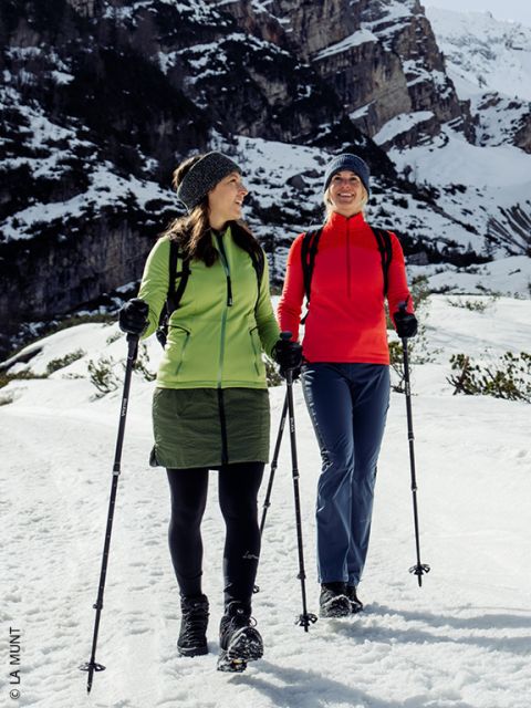 Zwei Frauen in Winterkleidung wandern mit Stöcken auf einem verschneiten Weg und lächeln sich vor dem Hintergrund schneebedeckter Berge an.