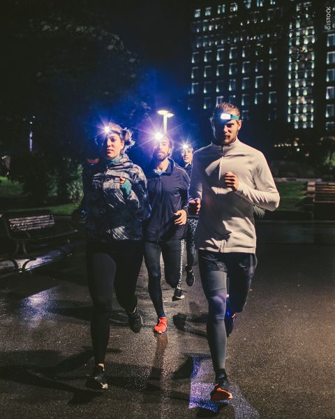 Vier Personen joggen nachts mit Stirnlampen auf einem schwach beleuchteten Weg. Im Hintergrund sind beleuchtete Gebäude zu sehen. Der Text lautet „Sicher & Sichtbar Laufen im Dunkeln“ und weist auf die Sichtbarkeit beim Laufen im Dunkeln hin.