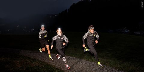 Zwei Personen laufen nachts mit Stirnlampen und orangefarbenen Jacken. Der Text lautet „SICHER & SICHTBAR SPORT IM DUNKELN“. Der Hintergrund ist eine verschwommene Stadtlandschaft mit Lichtern.