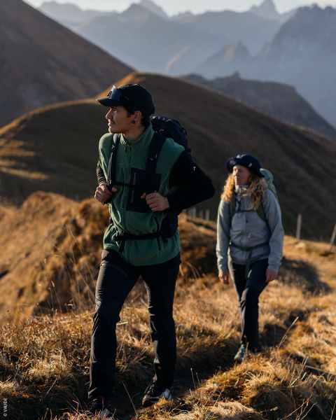 Zwei Wanderer in Outdoor-Ausrüstung gehen auf einem sonnenbeschienenen Bergpfad mit schroffen Gipfeln und grasbewachsenen Hügeln im Hintergrund.