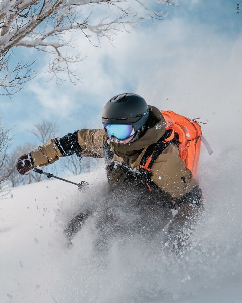 Ein Skifahrer mit Helm, Skibrille und orangefarbenem Rucksack fährt auf einem sonnigen, verschneiten Berghang durch tiefen Pulverschnee.