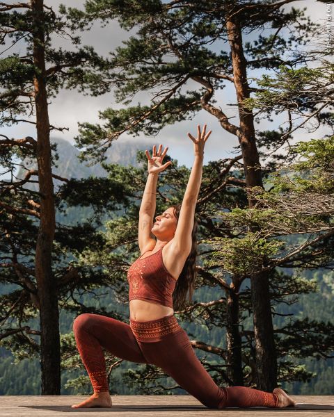 Eine Frau in roter Sportkleidung praktiziert Yoga im Freien auf einer Holzterrasse, umgeben von hohen Bäumen und Bergen, und streckt ihre Arme in einer Ausfallschritt-Pose über den Kopf.