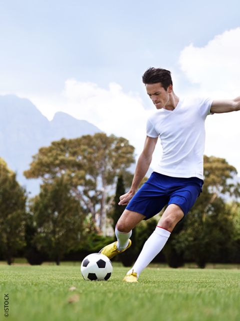 Ein Fußballspieler in einem weißen Hemd und blauen Shorts kickt einen Fußball auf einer Wiese mit Bäumen und Bergen im Hintergrund.