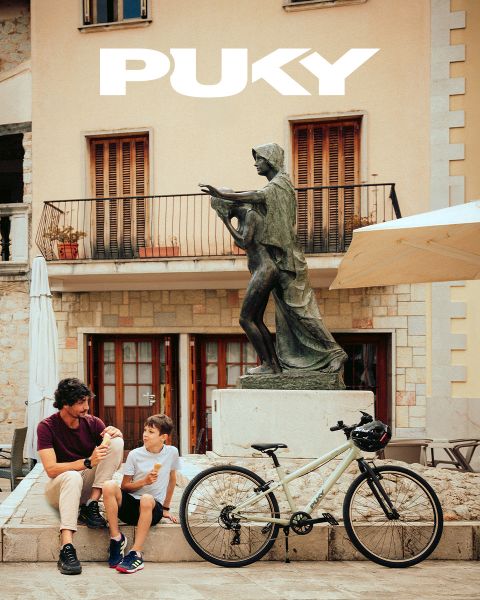 Ein junges Mädchen mit blauem Helm und gestreiftem Shirt fährt an einem sonnigen Tag auf einem hellblauen Fahrrad. Lächelnd fährt sie an bunten Graffitis in einer urbanen Umgebung vorbei. Das PUKY-Logo und der Slogan „Ready for life“ sind oben links zu sehen.