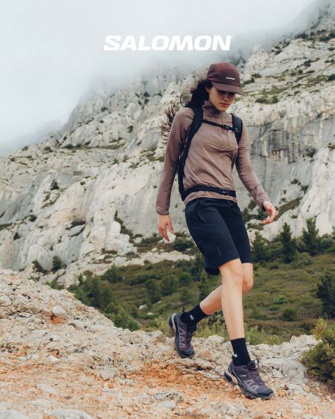 Nahaufnahme einer Person in Salomon-Schuhen und Salomon-Socken mit Farbverlauf in Blau. Sie steht auf einer Wiese mit einer malerischen Berglandschaft im Hintergrund. Ein Wanderer in Outdoor-Kleidung steht weiter entfernt, ebenfalls auf der Wiese.
