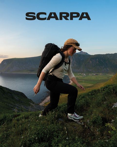 Eine Frau und ein Mann sitzen im Freien auf großen Felsen. Sie tragen Freizeitkleidung und Sonnenbrillen. Sie sind von Grün und Felsen umgeben, der Himmel ist leicht bewölkt. Oben im Bild ist das Wort „SCARPA“ zu sehen.