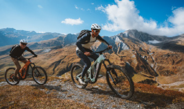 Zwei Radfahrer mit Helmen fahren Mountainbikes auf einer rauen Strecke mit malerischen Bergen und einem blauen Himmel im Hintergrund.