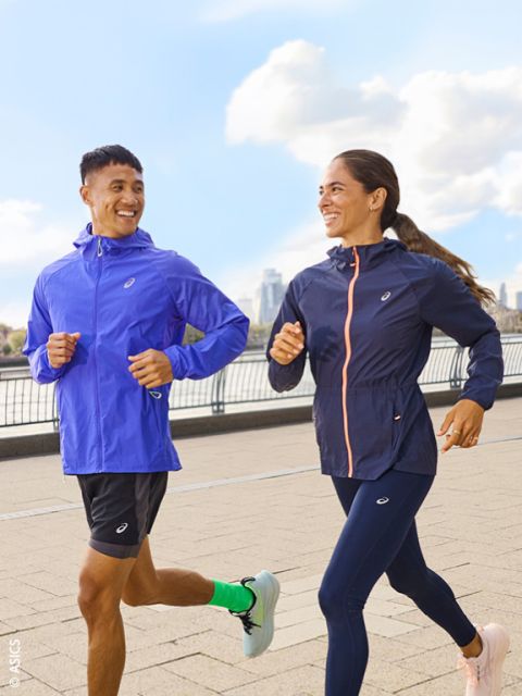 Zwei Menschen in Sportkleidung joggen im Freien auf einem gepflasterten Weg an einem Fluss und lächeln einander zu, während im Hintergrund die Skyline der Stadt und der blaue Himmel zu sehen sind.