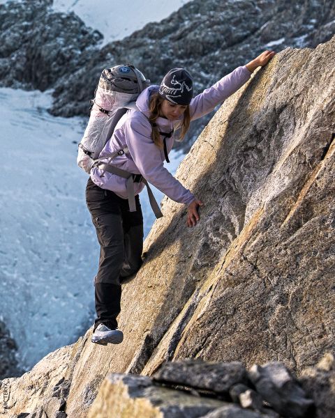 Eine Frau mit Rucksack klettert einen steilen, felsigen Berghang hinauf. Sie trägt eine Outdoor-Ausrüstung und einen Hut, während im Hintergrund schneebedeckte Gipfel zu sehen sind.