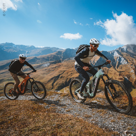 Zwei Radfahrer mit Helmen und Rucksäcken fahren mit ihren Mountainbikes auf einem felsigen Weg mit malerischen Bergen und blauem Himmel im Hintergrund.