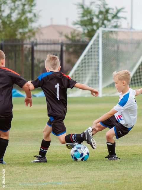 Drei Jungen spielen auf einer Wiese Fußball; einer in Weiß schießt den Ball, während zwei in Schwarz versuchen, ihn in der Nähe des Tores zu blocken.