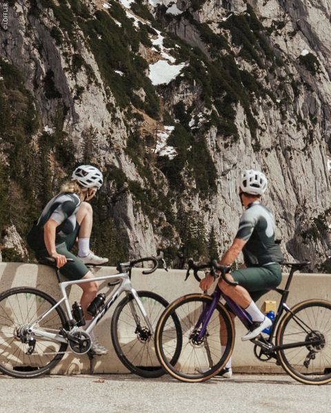 Zwei Radfahrer in passender Kleidung halten an einer Bergstraßenschranke an, mit felsigen Klippen und Schneeflecken im Hintergrund.