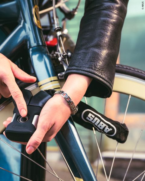 Eine Person in einer Lederjacke schließt ein blaues Fahrrad mit einem ABUS-Fahrradschloss ab, wobei sie sich auf die Sicherung des Rahmens und des Rads konzentriert.