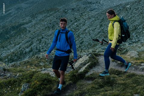 Zwei Personen in Outdoor-Kleidung wandern auf einem felsigen Bergpfad mit Trekkingstöcken durch das zerklüftete Gelände und die grüne Landschaft im Hintergrund.