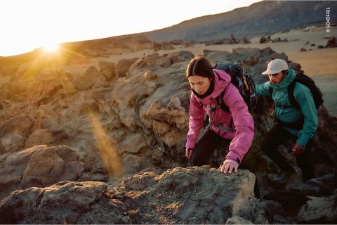 Zwei Wanderer in bunten Jacken klettern bei Sonnenaufgang über felsiges Gelände, während das Sonnenlicht über die zerklüftete Landschaft im Hintergrund fällt.