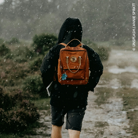 Eine Person, die eine Kapuzenjacke und einen orangefarbenen Rucksack trägt, geht auf einem regnerischen, schlammigen Weg, der von Grünflächen umgeben ist.