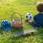 Kleiner Junge sitzt mit einem Fußball im Gras. In der Nähe stehen ein Korb mit bunten Eiern, ein blauer Helm und ein Skateboard. Sonnige grüne Landschaft.