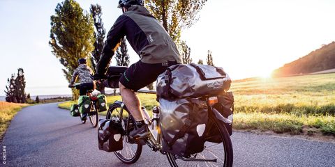 Ein Radfahrer fährt an einem klaren Tag Rennrad. Der Fahrer trägt einen Helm, eine Sonnenbrille und ein schwarzes Radsport-Outfit. Im Hintergrund ist eine Berglandschaft unter blauem Himmel zu sehen.