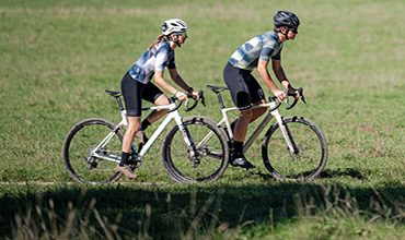 Ein Radfahrer fährt bei Sonnenuntergang auf einer asphaltierten Straße mit einer Berglandschaft im Hintergrund. Die Sonnenstrahlen erzeugen ein warmes Leuchten und der Radfahrer trägt einen Helm und eine Sonnenbrille.