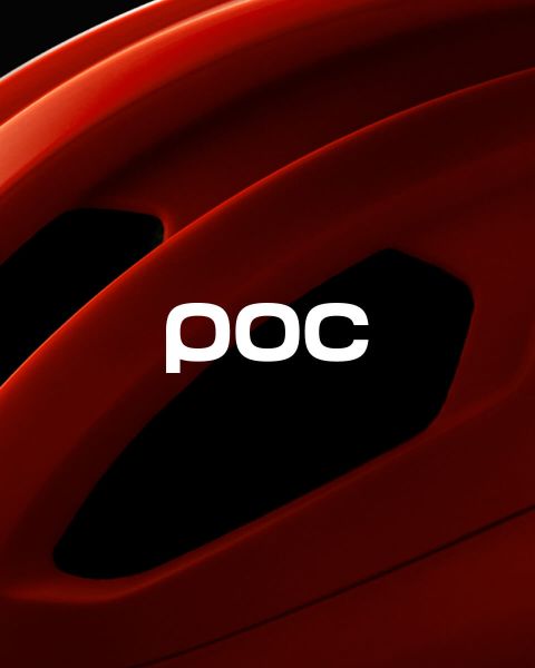 Eine Person in orangefarbenem Wintermantel und Helm steht auf einem schneebedeckten Berggipfel und blickt auf die fernen schneebedeckten Berge. Das POC-Logo ist auf dem Helm und in der oberen rechten Ecke des Bildes zu sehen.