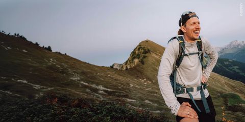 Ein Wanderer mit einem großen Rucksack und Trekkingstöcken steht auf einem felsigen Bergpfad unter einem strahlenden Himmel, umgeben von Wolken und fernen Gipfeln.