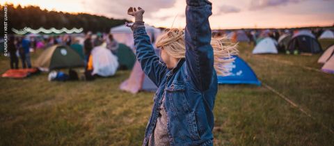 Eine Person in Jeansjacke mit erhobenen Armen steht bei Sonnenuntergang auf einer Wiese auf einem Campingplatz mit mehreren Zelten und Menschen.