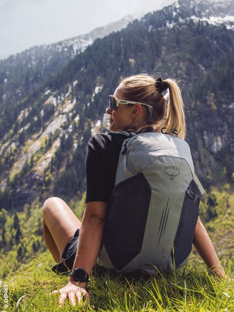 Frau mit Sonnenbrille und grauem Rucksack sitzt auf einem Grashügel und blickt auf eine bewaldete Berglandschaft in der Ferne.