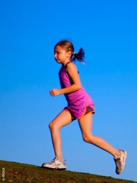 Junges Mädchen in einem rosafarbenen Sportoutfit, das im Freien auf einem Hügel mit einem klaren blauen Himmel im Hintergrund läuft.