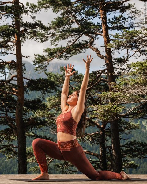 Eine Frau in roter Sportkleidung praktiziert Yoga im Freien auf einer Holzterrasse, umgeben von hohen Bäumen und Bergen, und streckt ihre Arme in einer Ausfallschritt-Pose über den Kopf.