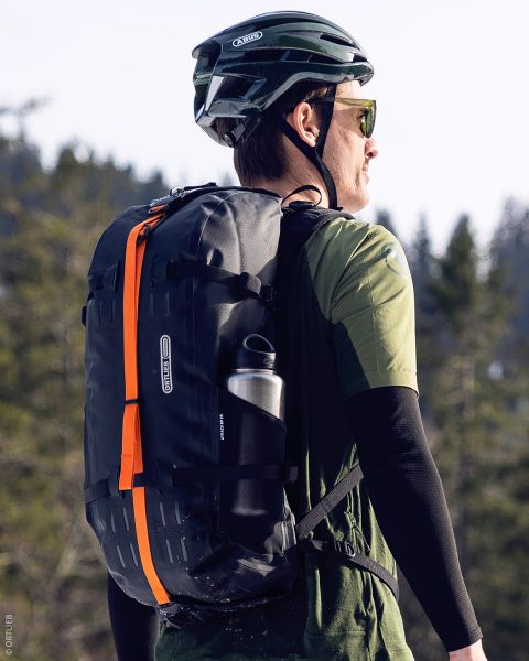 Eine Person, die einen grünen Helm und eine Sonnenbrille trägt, einen schwarzen Rucksack mit einem orangefarbenen Streifen und eine Wasserflasche mit sich führt, befindet sich im Freien in einem bewaldeten Gebiet.