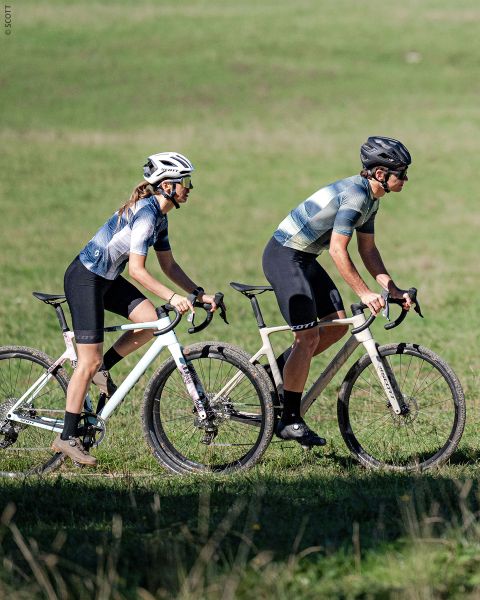 Zwei Radfahrer mit Helmen und Fahrradkleidung fahren mit weißen Fahrrädern nebeneinander auf einem grasbewachsenen Weg, mit einer grünen Wiese im Hintergrund.