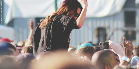 Eine Frau auf den Schultern eines Mannes genießt ein Konzert im Freien, umgeben von einer Menge, die Fotos macht und die Hände hebt.