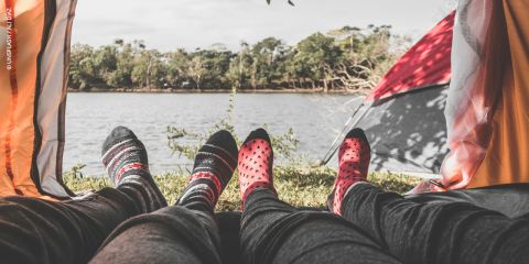 Zwei Personen, die gemusterte Socken tragen, entspannen sich in einem Zelt, mit Blick auf einen See und Bäume draußen an einem hellen, ruhigen Tag.