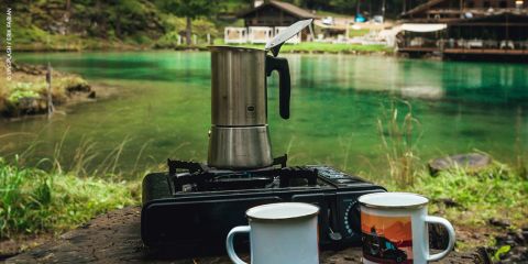 Eine Mokkakanne auf einem Campingkocher mit zwei Bechern davor, auf einem Tisch an einem türkisfarbenen See mit Holzhütten im Hintergrund.
