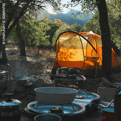 Ein leuchtend orangefarbenes Zelt auf einem bewaldeten Campingplatz mit Kochgeschirr und Campingzubehör im Vordergrund.