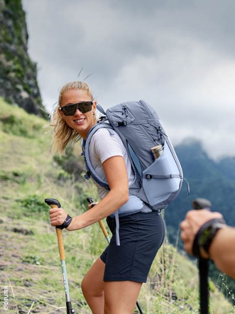 Eine Frau mit Sonnenbrille wandert mit Trekkingstöcken und einem grauen Rucksack einen grasbewachsenen Bergpfad hinauf und lächelt in die Kamera.