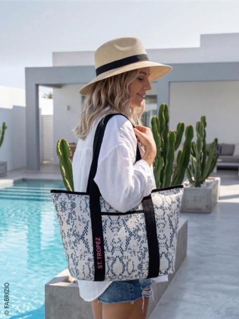 Eine Frau mit Strohhut und weißem Hemd steht an einem Pool und hält eine große gemusterte Tragetasche mit der Aufschrift "St. Tropez" in der Hand, mit Kakteen im Hintergrund.
