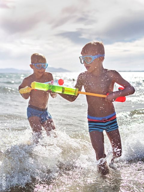 Zwei Jungen mit Schwimmbrillen spielen mit einer Wasserpistole im Meer und planschen in den Wellen unter einem hellen, sonnigen Himmel.