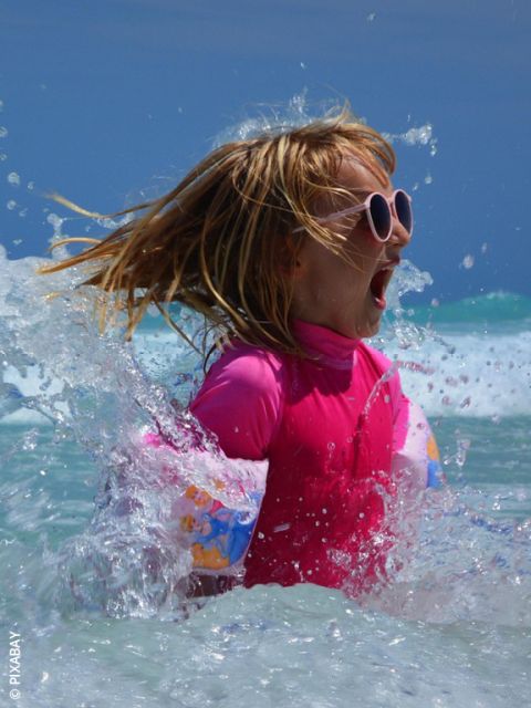 Ein Mädchen mit rosafarbener Sonnenbrille und Badeanzug plantscht im Meer und lächelt aufgeregt, als das Wasser an einem sonnigen Tag um sie herum spritzt.