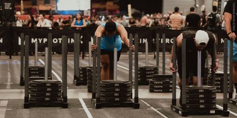 Bei einem gut besuchten Indoor-Fitnesswettbewerb schieben zwei Sportler mit Gewichten beladene Schlitten. Im Hintergrund beobachten Zuschauer das Geschehen hinter Absperrungen.