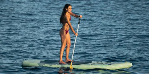 Eine Frau im Bikini steht auf einem Paddleboard, hält ein Paddel in der Hand und lächelt auf dem ruhigen blauen Wasser.