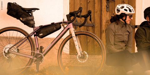 Zwei Radfahrer, die Helme und Jacken tragen, sitzen auf dem Boden in der Nähe einer Mauer; neben ihnen steht ein voll beladenes Tourenrad.