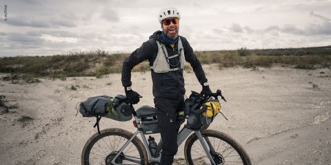 Ein lächelnder Radfahrer mit Helm und Sonnenbrille steht mit einem voll beladenen Tourenrad in einer sandigen, bewölkten Landschaft.
